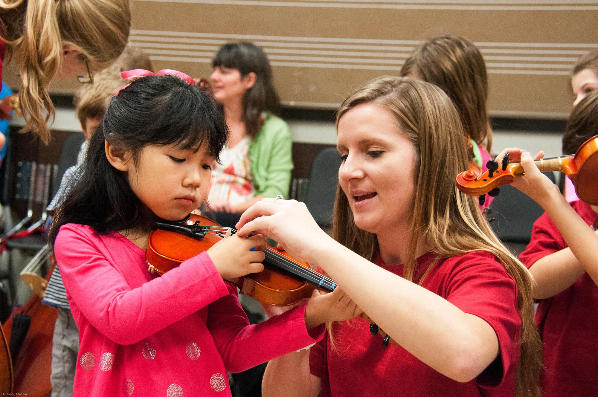 a violin teacher helps a very young student with hand position in a group violin class.