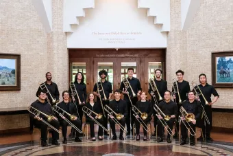 A group of students dressed in black holding trombones and posing in two rows in front of the doors to the John and Nellie Connelly Banquet Hall.