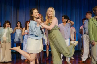 Two singers clasp hands as they perform a show number on stage, while other singers watch in the background.